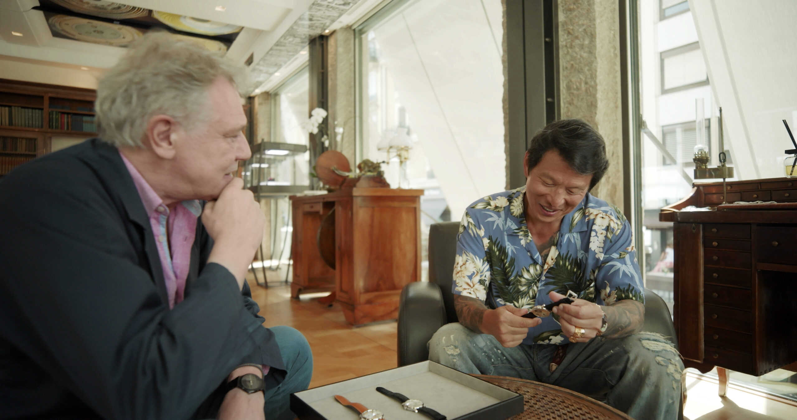 Wei Koh and François-Paul Journe sitting together indoors, smiling as they examine watches during a Man of the Hour interview scene