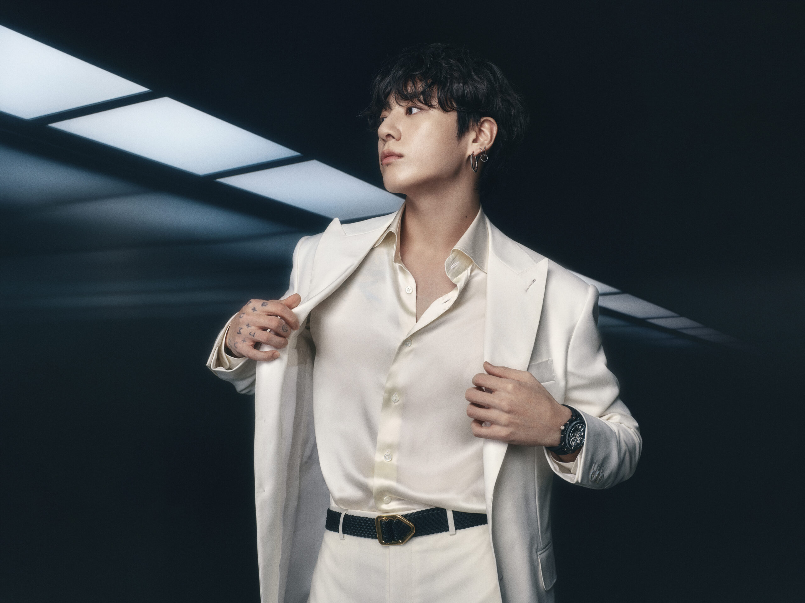 Jung Kook wearing a white suit and Hublot's Big Bang Original Unico Black Magic watch in a studio setting
