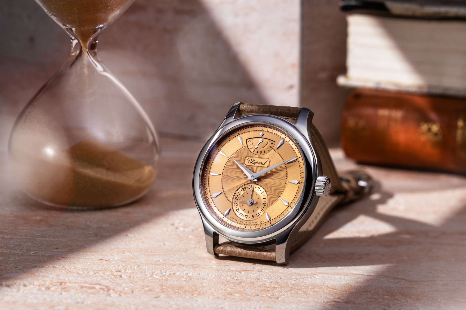 The Chopard L.U.C Quattro Origin leaning against a stone surface with a half-filled hourglass to its left and stacked vintage books behind, bathed in warm directional sunlight