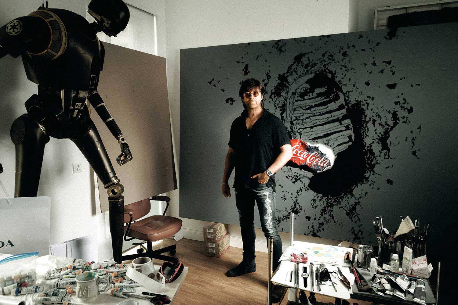 Michael Moebius in his Dresden studio. Behind him is a new work titled First Trash, reflecting on the contrast between human progress and the traces we leave behind. To his right is a mechanical robot sculpture carrying luxury shopping bags (©Revolution / Photography René Gaens)